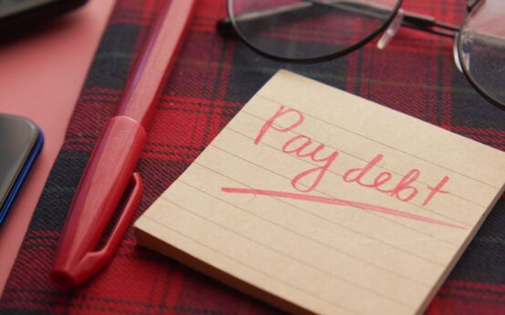 Close-up of a note reading 'Pay debt' next to a red pen on a plaid fabric, emphasizing financial reminders.
